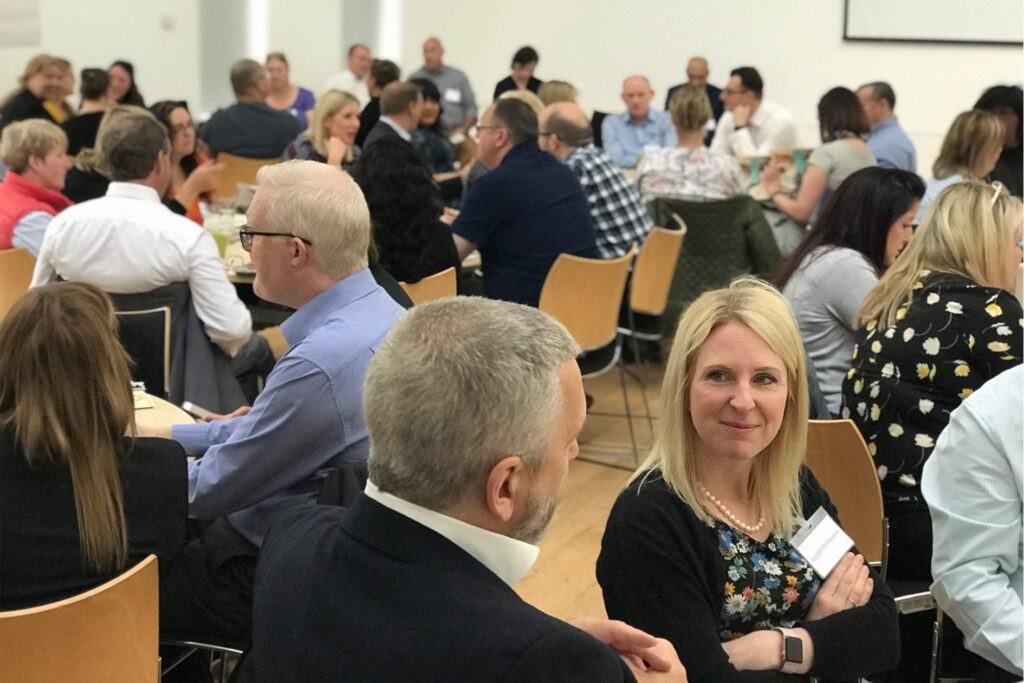 A large room is full of people at multiple tables. They are sitting down talking to other people at their tables.