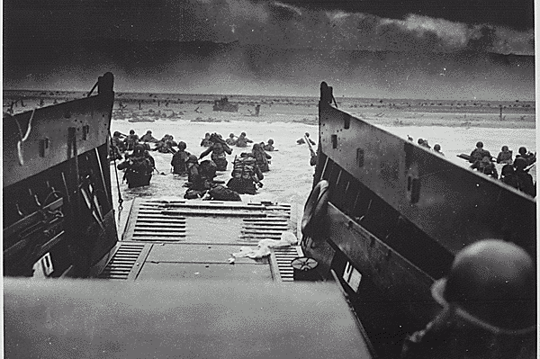 An original D-Day photo, taken from the inside of a landing craft. The black and white image shows a landing craft with the ramp down and soldiers wading through the sea towards the beach.