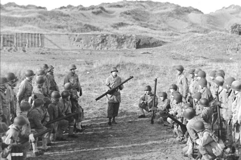 A black and white image of US troops armed with bazookas. They are gathered in two groups, one on each side of the picture, all looking towards one man in the middle holding a bazooka. Behind them is the concrete wall at Braunton Burrows.