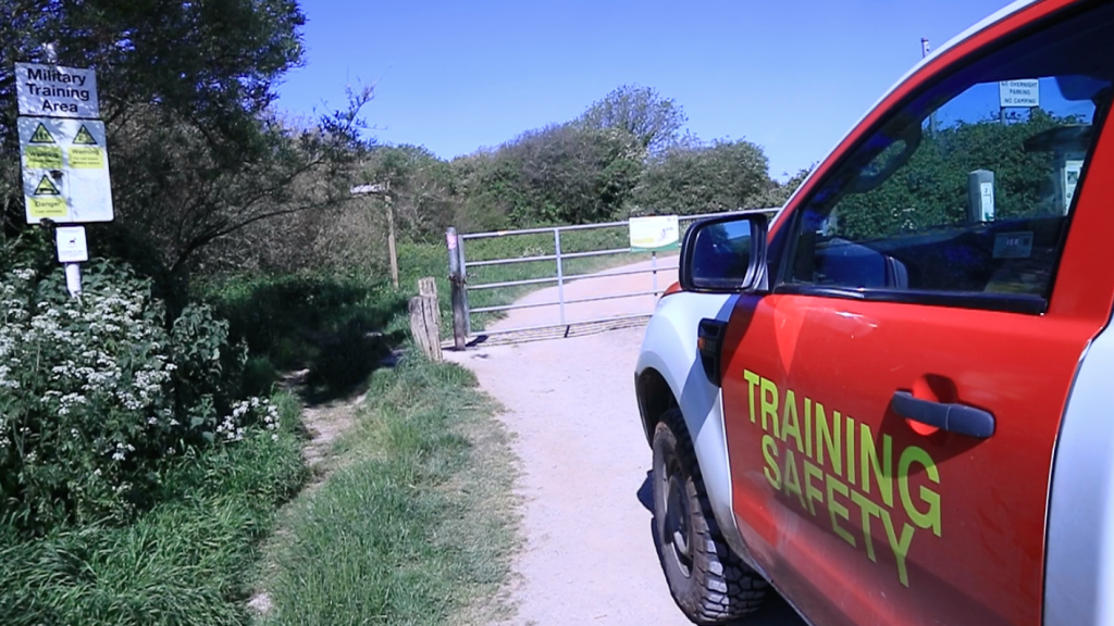 A modern Training Safety vehicle with red and white livery on the right of the image, parked in front of a gate. On the left is a sign warning of military training.