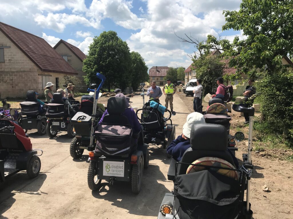 A DIO archaeologist is standing at the front of Copehill Down Village, an urban training area on the history of the site. The village consists of purpose fit urban houses and facilities used to train soldiers. The Disabled Ramblers are in a group listening to the talk.