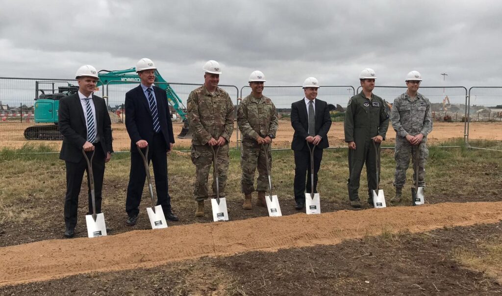 7 men including representatives from DIO and Kier VolkerFitzPatrick hold spades are in white helmets standing next to a line of sand for the ground breaking ceremony at RAF Lakenheath.