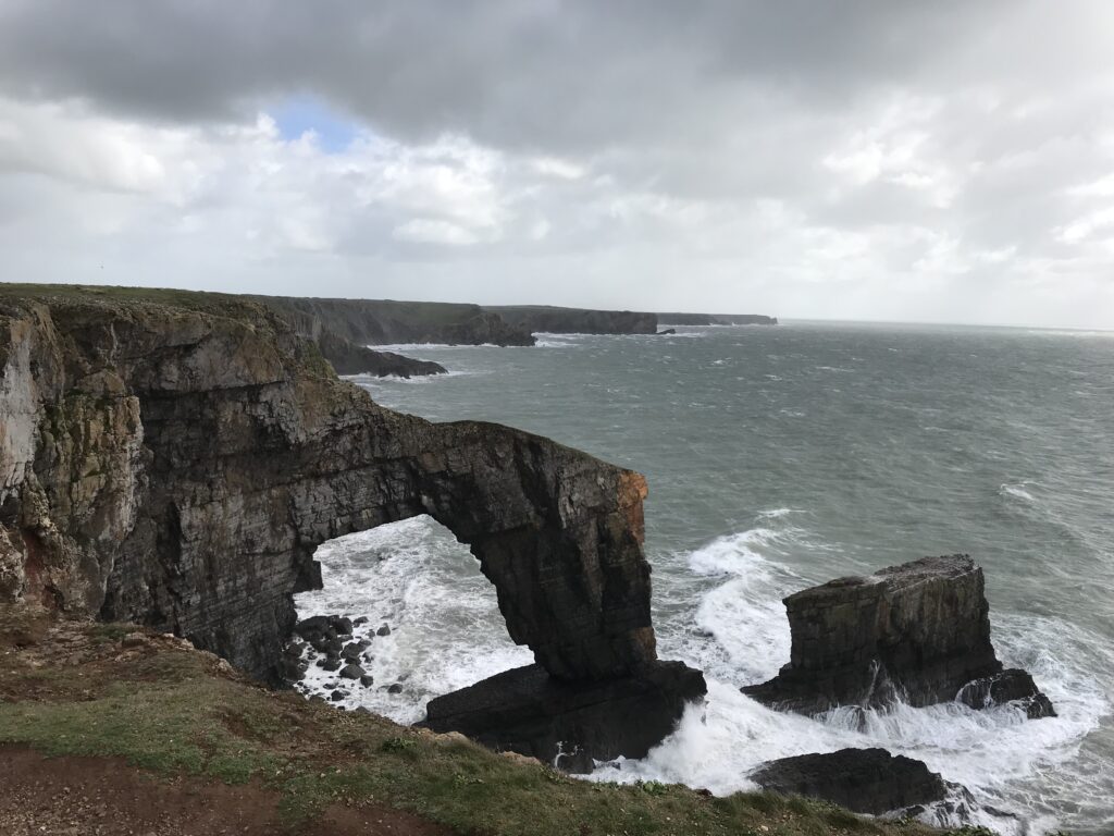 A picture of a the green bridge of Wales at the Castlemartin Ranges. There is sea overlooking the bridge.