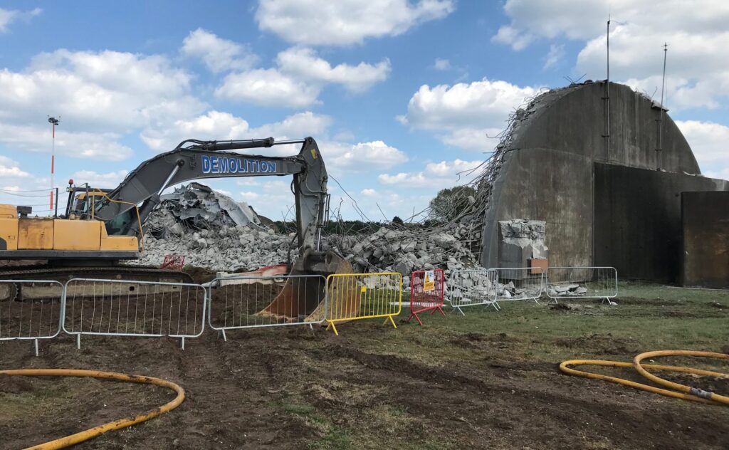 A hangar at RAF Lakenheath is being demolished with a vehicle. There are materials from the demolished hangar and rubble pictured next to the remaining building.