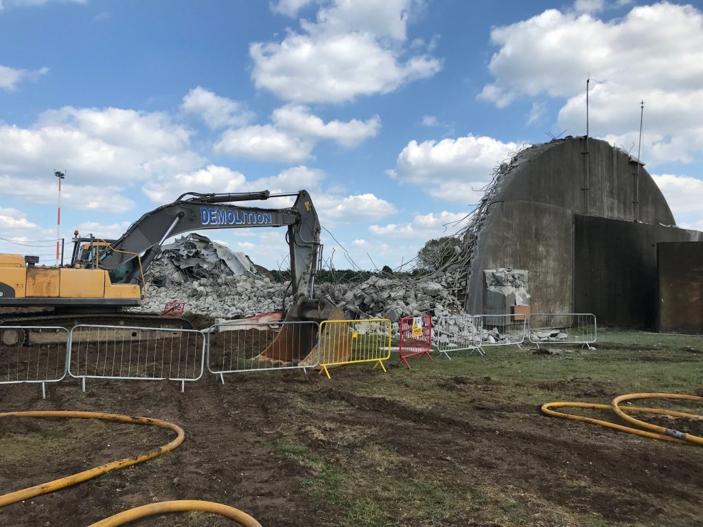 A yellow crane on the left is demolishing a grey hangar on the right. There is bricks and rubble next to the demolished building.