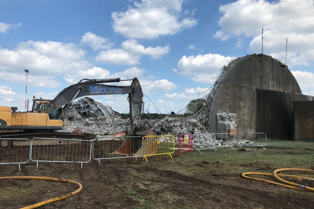 A large digger behind a fence, with a pile of rubble and a remaining semi-circular concrete wall which still stands. 