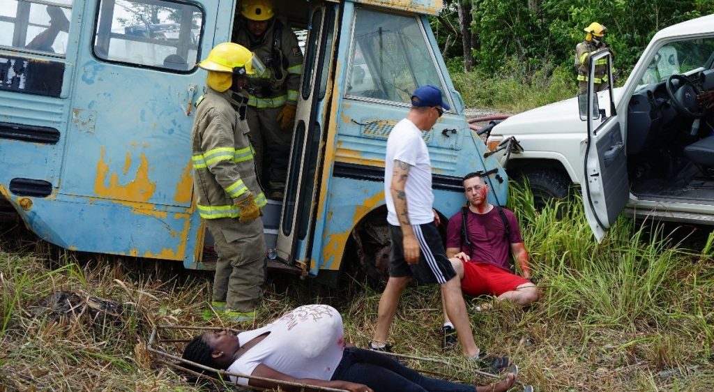 Pictured is a light blue tourist bus with two members of the Belize Defence Force next to it. A casulty is infront of the bus and another casulty is lying down on grass. A vehicle is also pictured next to the bus.