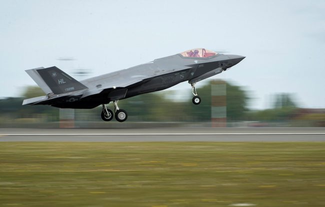 Pictured is an F35 taking off on a runway. The F35 is grey with 3 wheels. There is a little window at the front where you can see the pilot. There is grass next to the runway