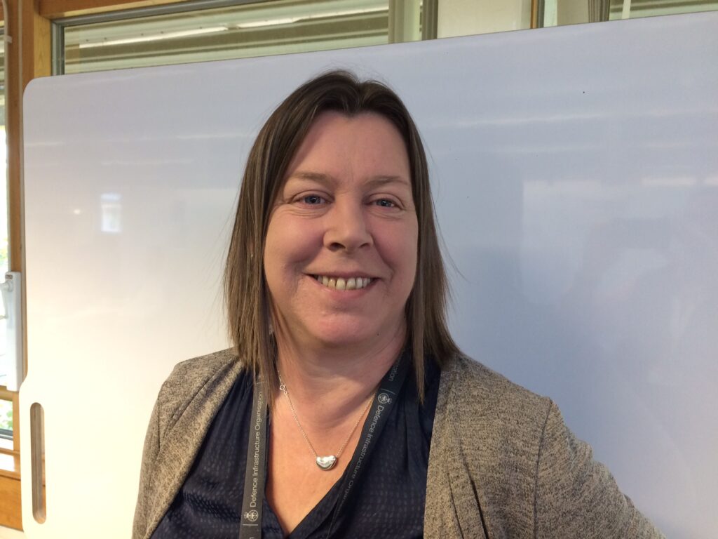 Claire is pictured infront of a white board. She has brown hair and is wearing a black shirt with brown cardigan