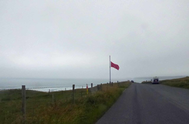 A red flag is located on some grass and fencing near a road. There is sea behind the grass.