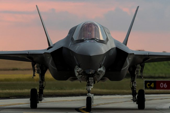 Pictured is an F35 Lightning jet. The Jet is facing the front and is grey with a window at the front and 3 wheels, one at the front and two at the back.It has two grey wings at the back and is pictured on a road with a field behind it. The sky is pink/blue.