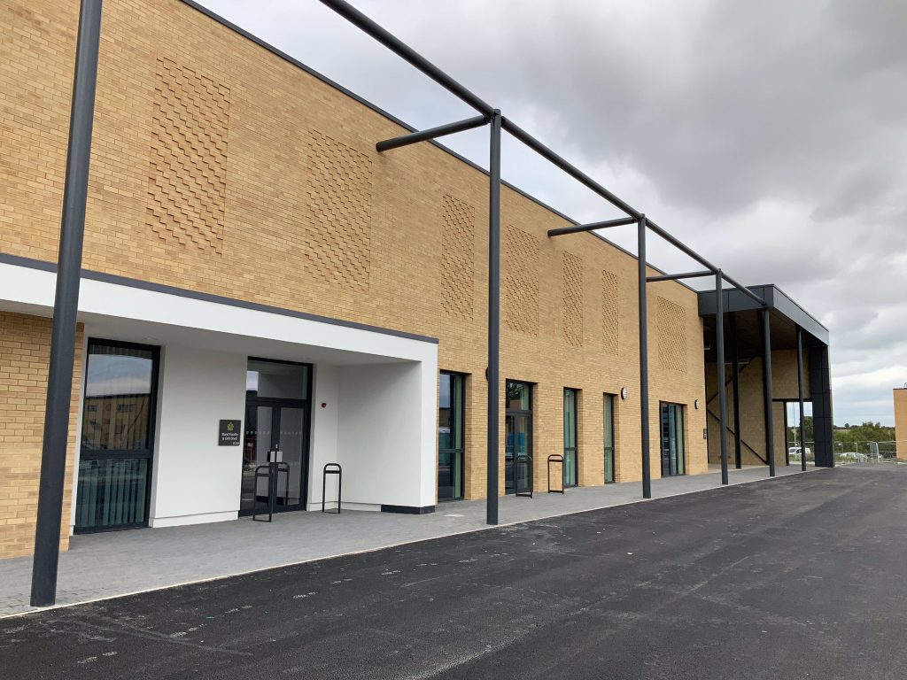 The HQ Regimental building is rectangle with bricks layered across. On the left there is a two black doors with clear windows for the entrance. Alongside this are a number of long windows. The building is black tarmac infront of it.