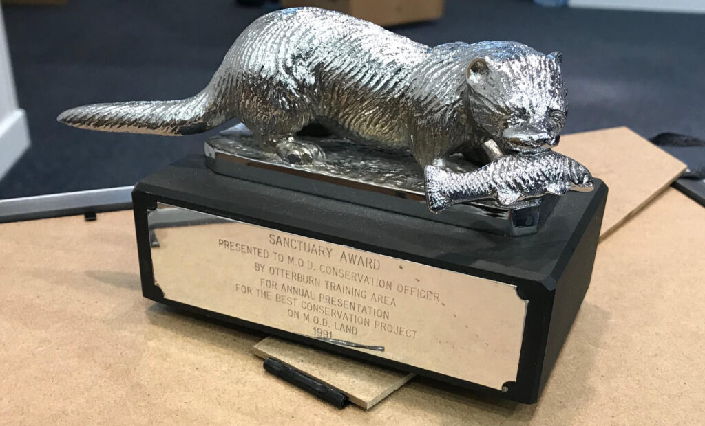 Picture of a silver and black award. The award is a black block with silver writing stating the Sanctuary Award and the top of the block has a Silver Otter on it with a silver fish in it's mouth.