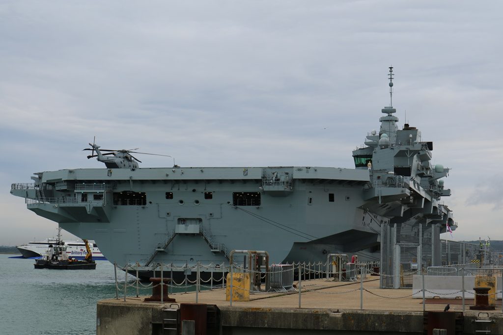 An image of HMS Queen Elizabeth, looking towards the stern, taken from a jetty and showing the ship alongside another jetty. 