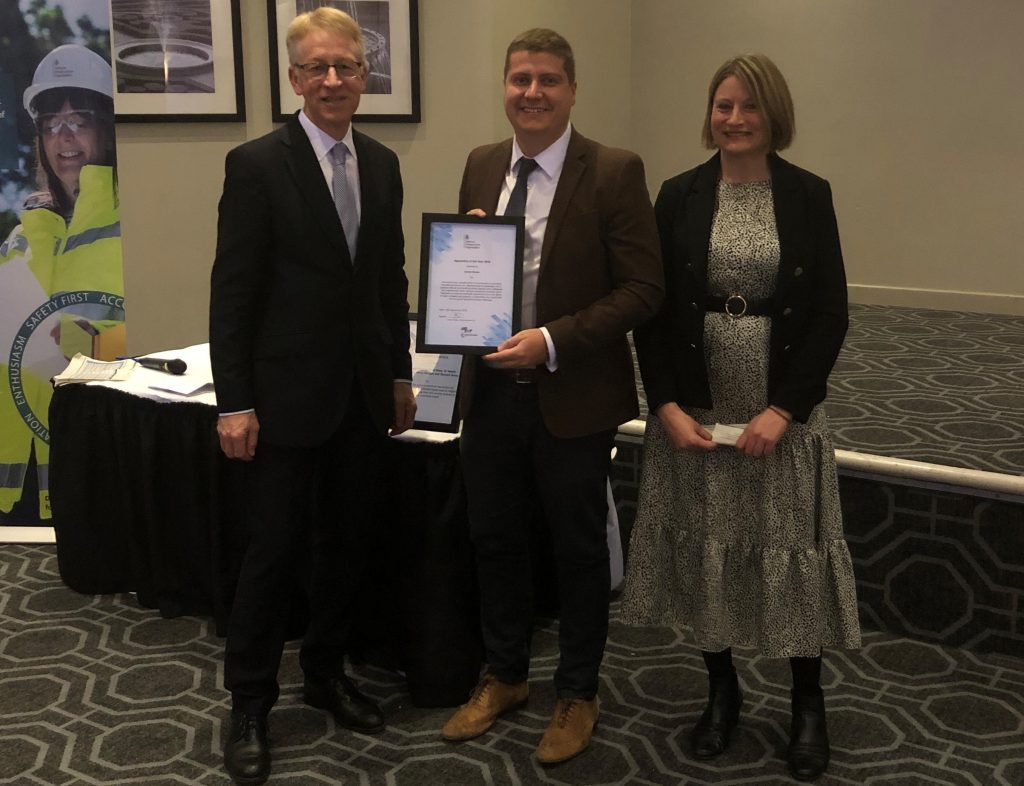 Pictured is James Davies, Apprentice of the Year in the middle holding up his framed certificate award. He is wearing a brown suit with a white shirt and black tie. Pictured on his left is CEO of DIO Graham Dalton in a black suit and grey tie. Pictured on his right is DIO's head of communications Emma Reynolds, in a greay dress and black blazer.