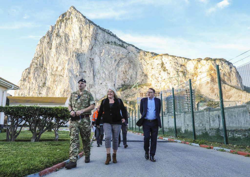 Jacqui Rock is walking alongside a soldier in uniform and a man in a suit. Behind her is the Gibraltar rock.