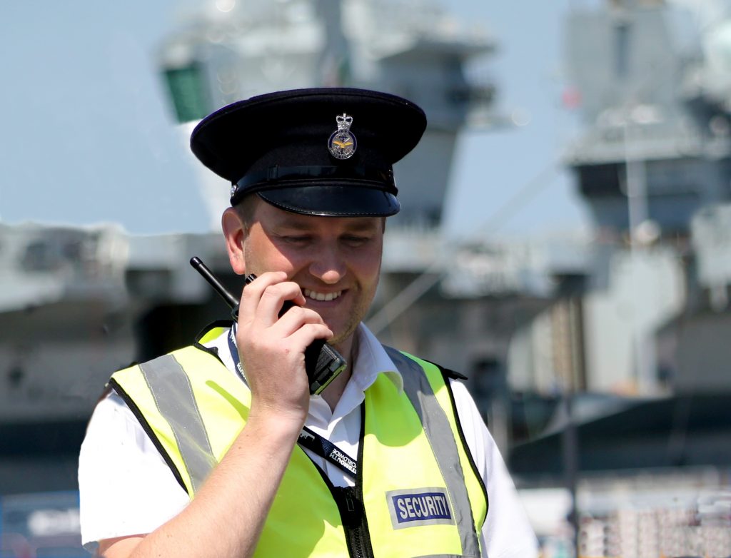 An MGS guard in uniform at Portsmouth naval base, using his walkie-talkie and with an aircraft carrier behind him.