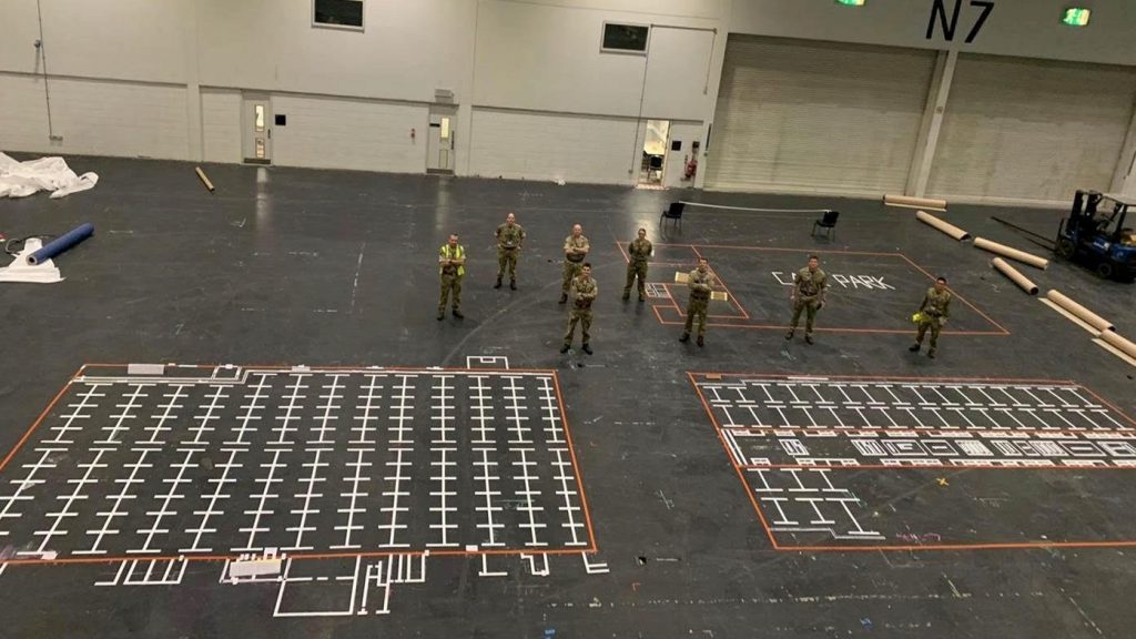 A mock up plan on the black floor drawn in white and red shows how the Nightingale Hospital may look. There is a box with a red outline and inside are lines with white marks and sections for where beds will go. There are some reservists standing near it looking at the plan.