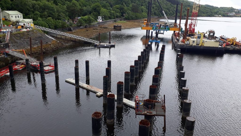 Pictured are a number of vertical brown cylinder piles that have been placed into the seabed. Pictured on the left is a white building with a silver bridge leading down to a boat.