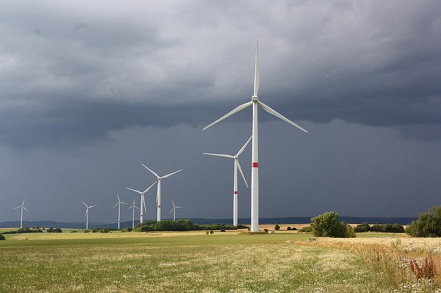 Pictured is 9 wind turbines behind each other on a field. The sky is cloudy.