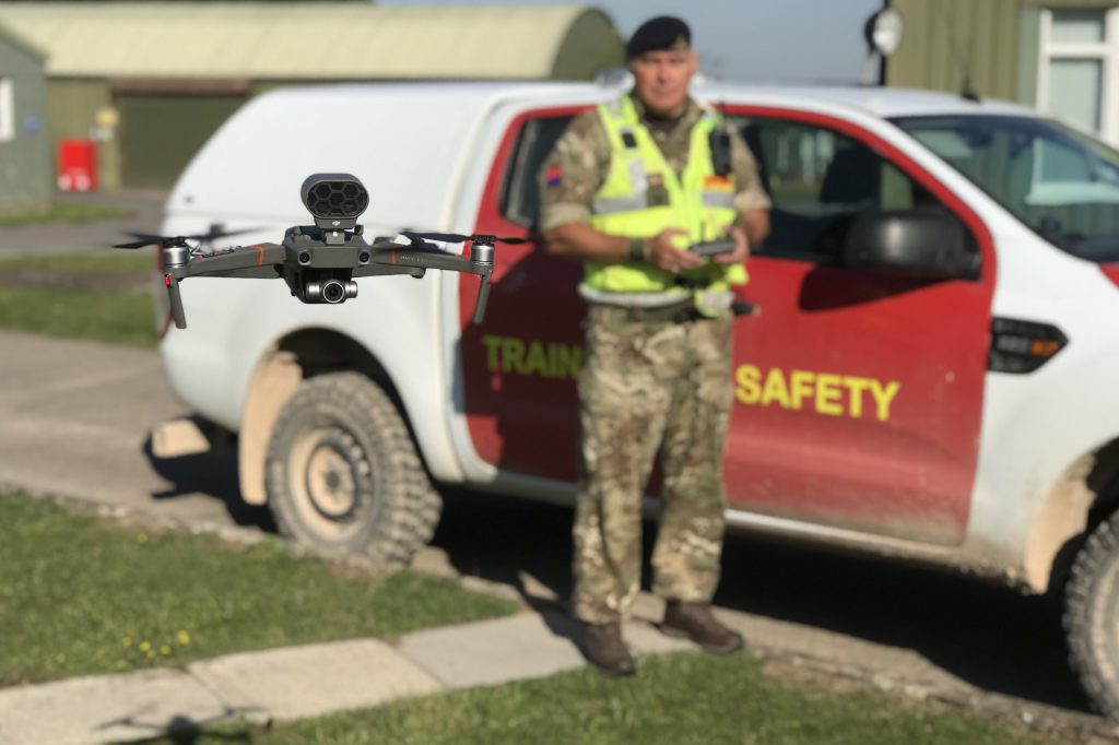 A Training Safety Marshal, wearing camoflage uniform and a high-vis vest, stands in front of a vehicle marked Training Safety. In his hands he holds a controller and near to the camera is a drone, with horizontal propellers on either side. The drone is in focus with the Training Safety Marshal, vehicle and background slightly out of focus.