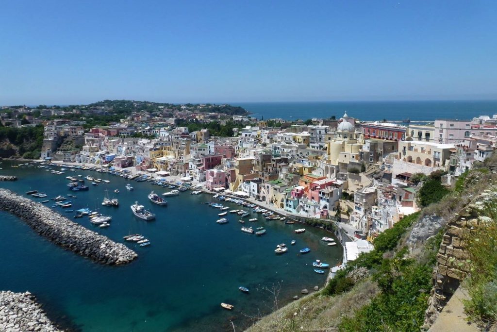 View of a marina and associated buildings and boats on a sunny day. The image is taken from a hill overlooking the marina.