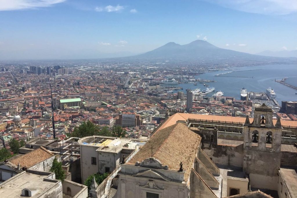 A view of Naples from a high point, with the rooftop of what appears to be a church in the foreground. Spread out behind are the rooftops of the city and to the right, the Bay of Naples with a number of cruise ships. In the distance is Mt Vesuvius.