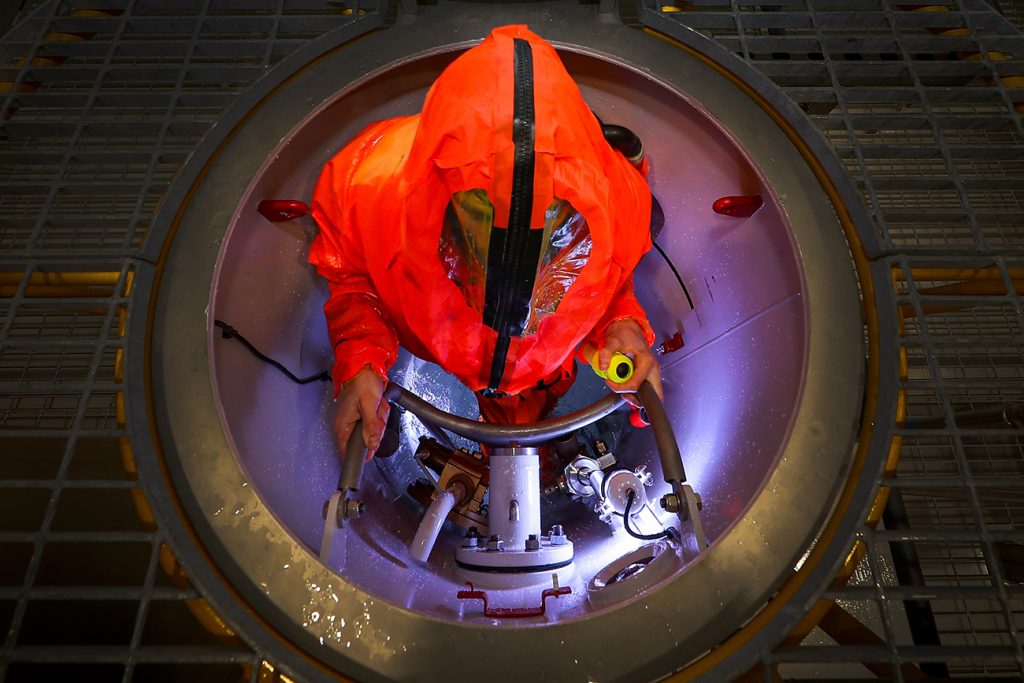 A sailor in an orange dry suit is standing in a round hatch, holding a metal ladder. The picture is taken from above and only their upper body is visible. 