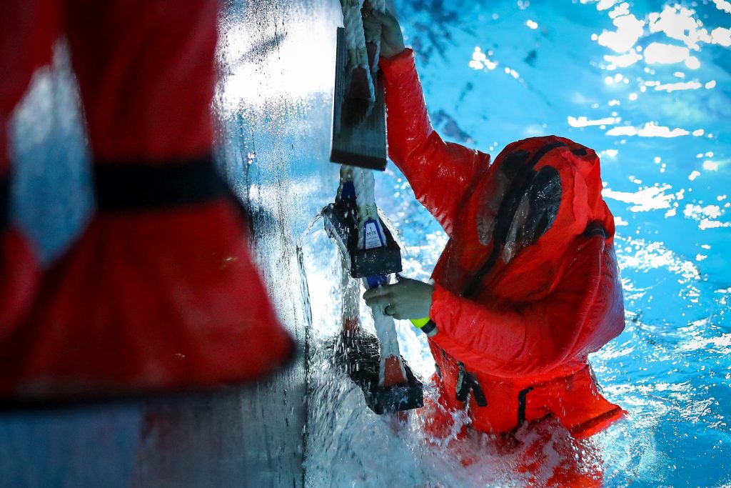 The left side of the image is a vertical wall. The right is a pool, with a sailor in an orange dry suit who is mostly in the water, but hanging on to a rope ladder with his hands.