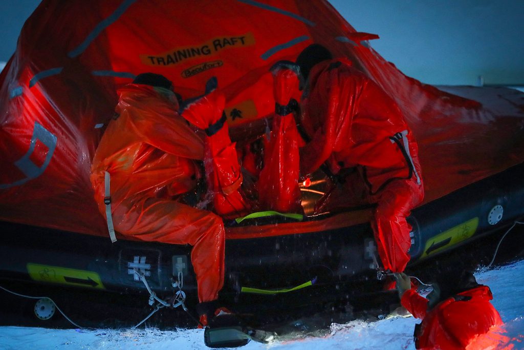 Two sailors in orange dry suits sit on the edge of an orange and black training life raft in a pool. 