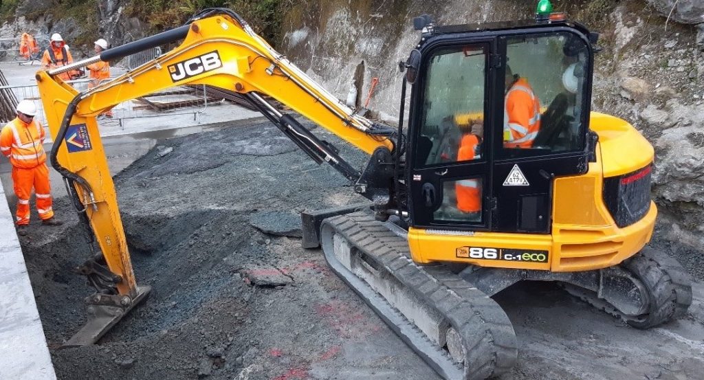 A yellow crane is pulling concrete out from the soil. There is a working in orange visible jacket sitting inside and some workers outside.