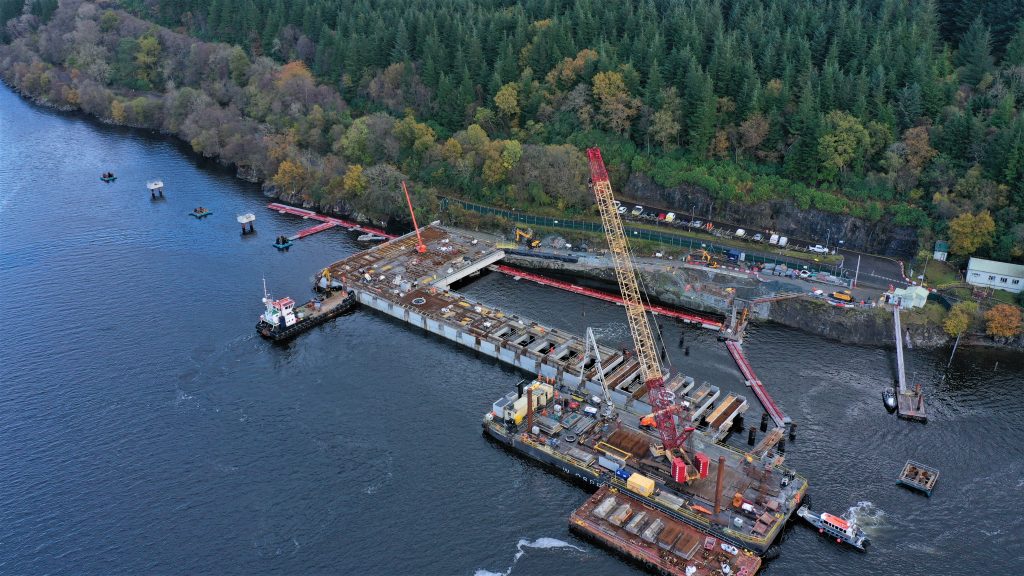 A drone image of the Northern Ammunition Jetty over the Loch Lomand sea. A big yellow and red crane is on the jetty along with a small white boat beside it. There is a road and lots of green trees on the right of the jetty.