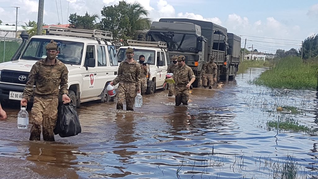 Six soldiers are walking through flooding with water cartons and bags. Next to them are white vehicles and a military truck.