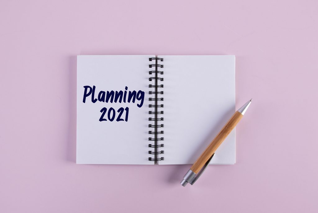 A notebook lies open. On the left hand page it reads "Planning 2021". The right hand page is blank but has a pen resting on it. The background is pink.