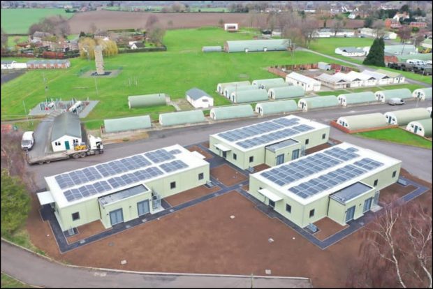 Aerial view of Nesscliffe Training Camp. In the foreground is an area of bare earth with three single storey buildings with solar panels on their roofs. In the background are multiple Nissen huts and a large area of grass.