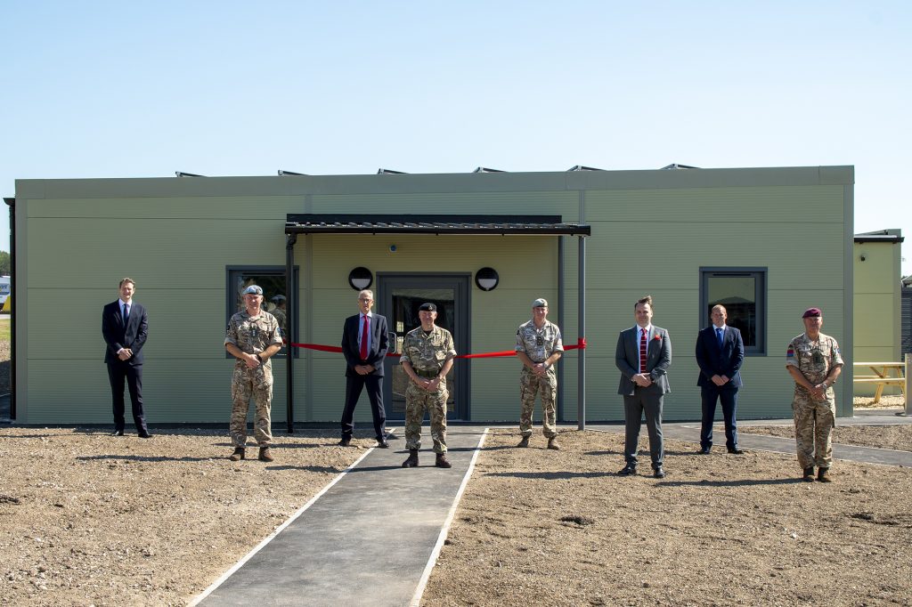 Eight men stand, socially distanced, outside a single storey green building. The foreground is bare earth, unlandscaped, with a path leading to the building. Four of the men are wearing military camouflage uniform, the others are dressed in dark suits.