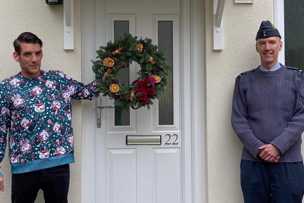 A white man in a Christmas jumper stands to one side of a front door. He holds a Christmas wreath. On the other side of the closed door is another white man in an RAF officer uniform. 