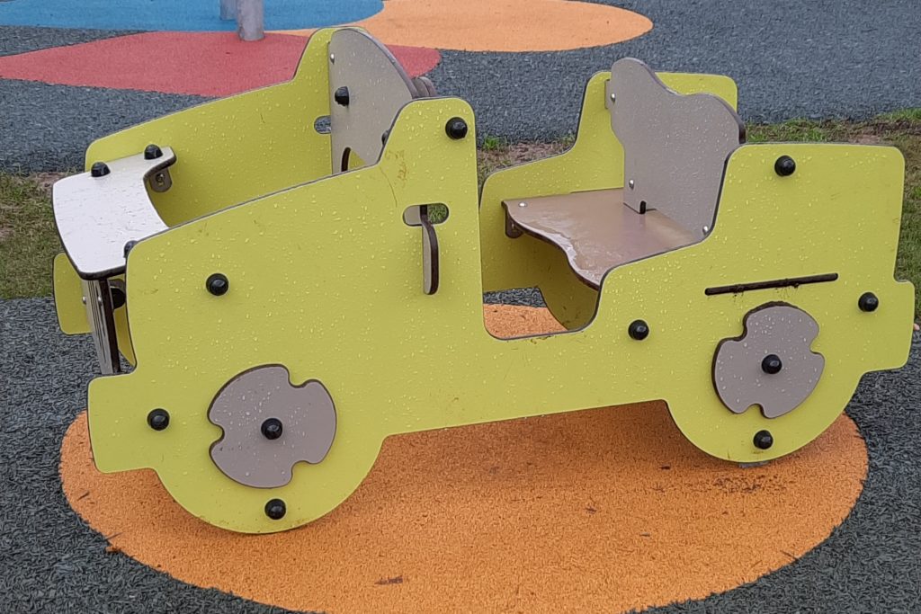 A car toy which children can sit inside on a playground. The car sits on an orange circle on the surface of the play park.