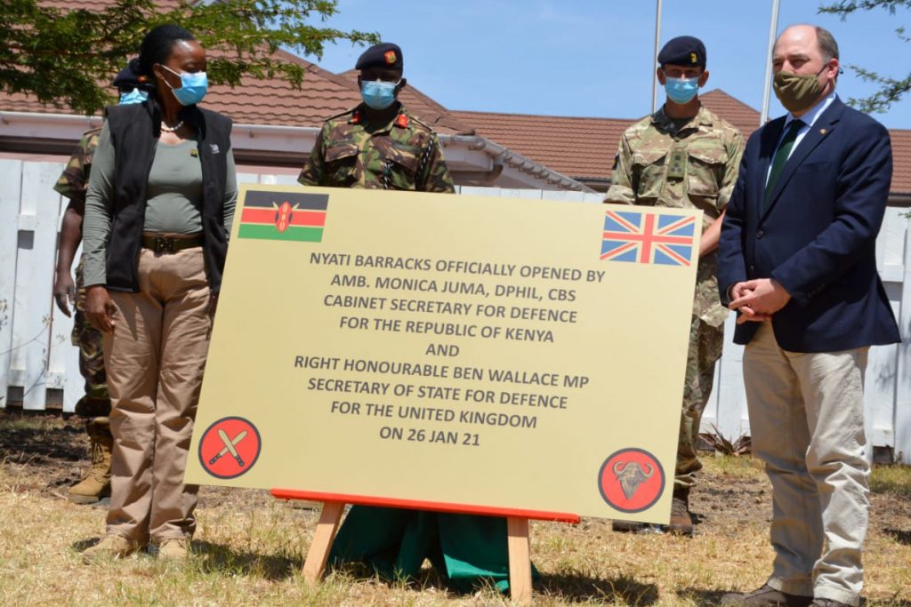 Ambassador Dr Monica Juma and Ben Wallace MP stand on either side of a large sign marking the opening of Nyati Barracks, with two uniformed Kenyan soldiers and one uniformed British soldier behind them. 
