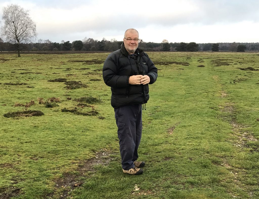 Peter is wearing a black puffed coat and black heans with brown trainers. He is wearing glasses and is standing on green heath at the Stanford Training Area. Behind him in the distance is a tree.