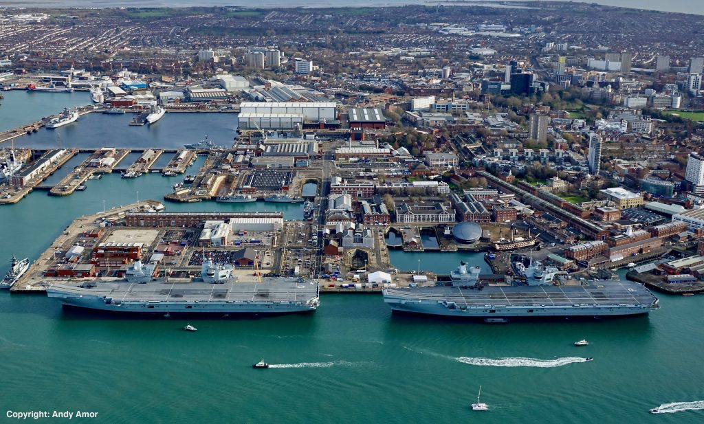 Both the Royal Navy's Queen Elizabeth Class aircraft Carriers pictured next to each other at the HMNB Portsmouth harbour. The water is a green/blue colour and there are buildings located behind the vessels.