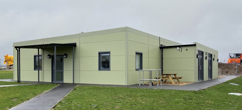 Light Green one floor modular building with two windows with grey boarding around it at the front and a dark green door. On the side is a wooden bench and around the building is grass. Located infront of the door is a grey path.