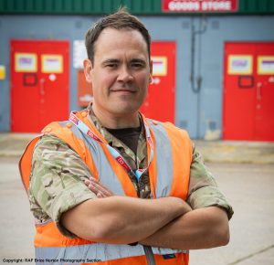 Seargent Ryan Duffy is wearing a sleeveless orange hi-vis top with a short-sleeved army shirt under it and has brown hair. He is standing infront of a grey building with 3 red doors.