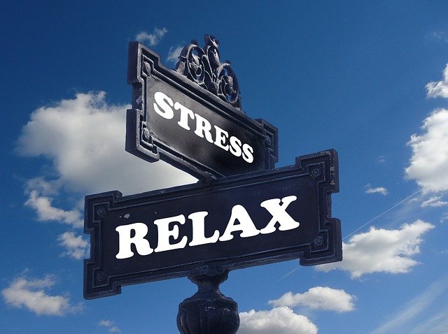 A black sign with white wording that say stress on it with another black sign under it that says relax in white text. There is sky and clouds behind the sign.