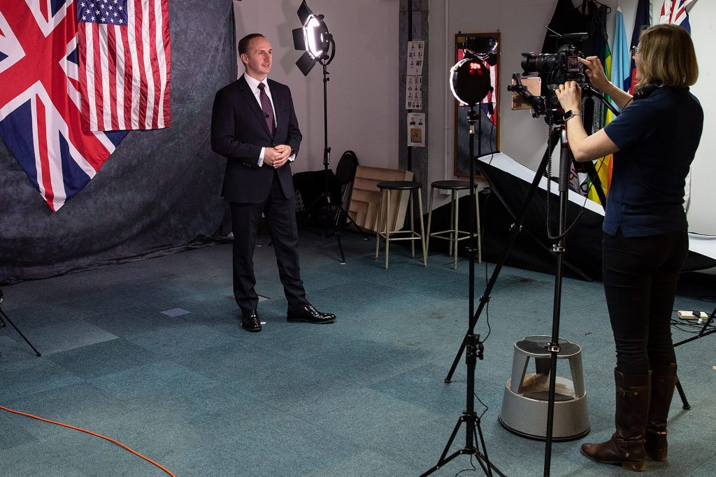 A white man in a suit stands in front of an American flag and a Union flag. He is looking at a woman holding a camera.