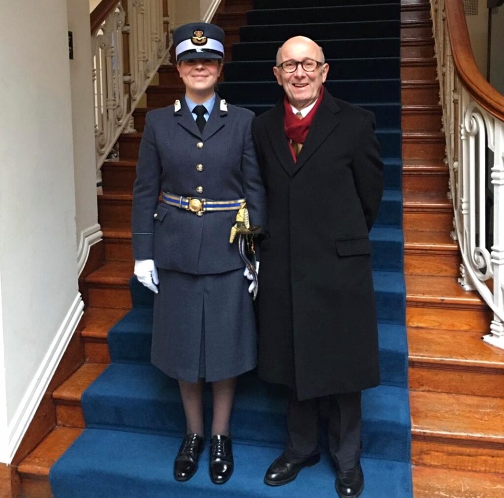 Annabel is standing on some stairs with a dark blue carpet wearing a long navy blazer jacket with three gold buttons and a gold and blue belt around it. She has a long navy skirt on and black shoes. She is also wearing a hat with the Royal Air Force symbol in circle in the middle. Next to her is her father in a long black coat, black shoes and black glasses.