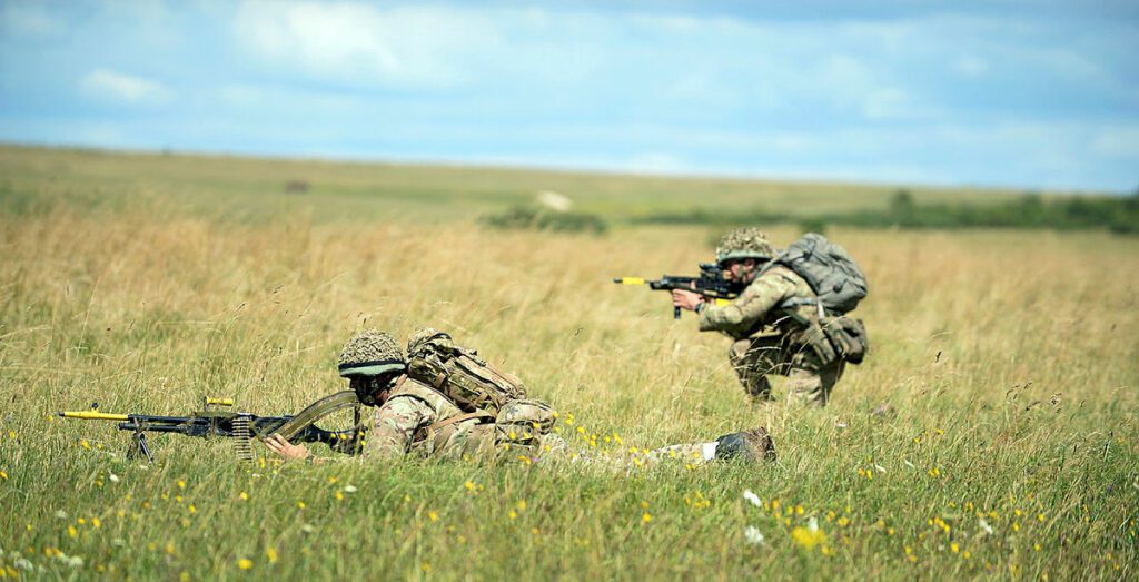 A soldier is lying down on some grass with a rifle another soldier is kneeling with his rifle. They are surrounded in grass and it is a sunny day.