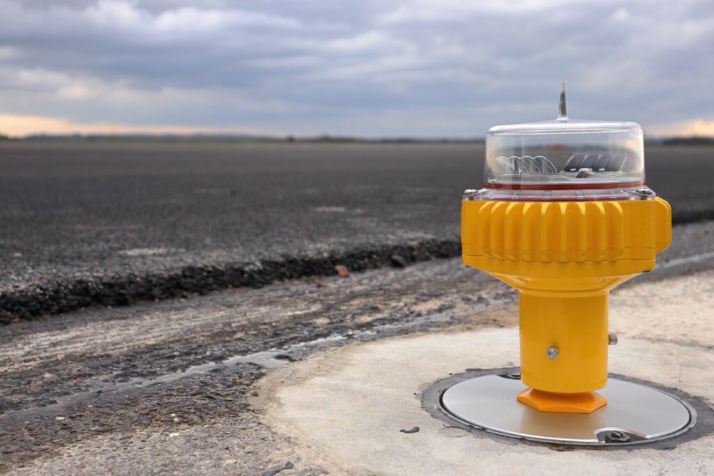 In the foreground is a light and in the background is the resurfaced runway. The light is a yellow object emerging from the ground. It flares wider at the top with a clear covering.