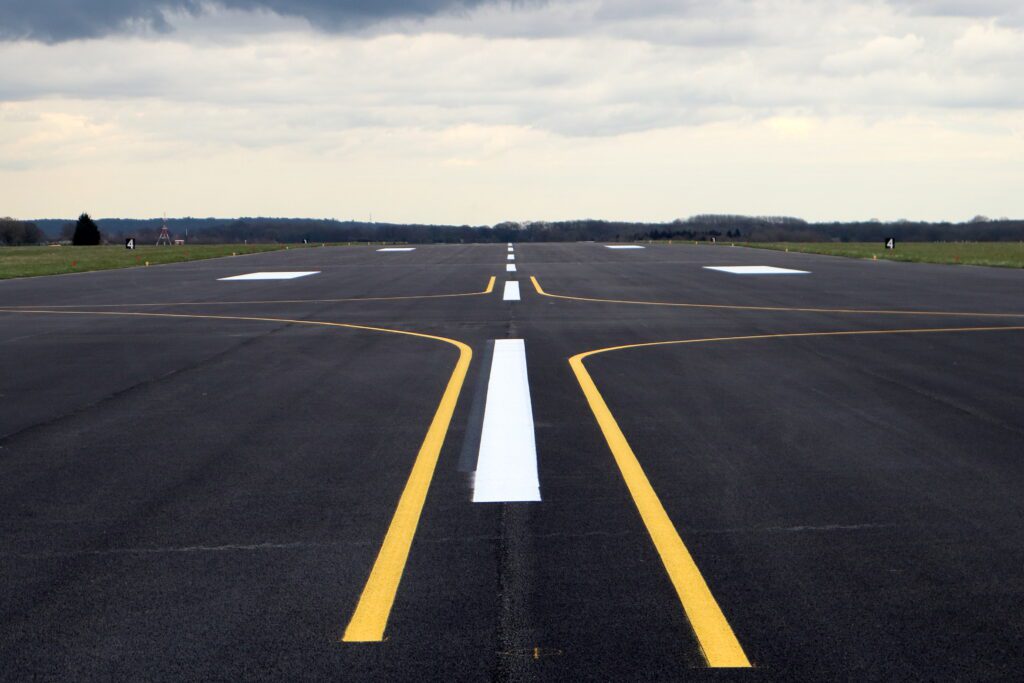 The foreground is the resurfaced runway. The surface is dark and has a dotted white line progressing up the middle with yellow lines arcing away to the each side.