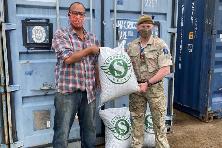 Two men stand in front of a couple of shipping containers. On the left is a black man, dressed casually, and on the right is a white man dressed in military uniform. Both are facing the camera and holding a sack of seedballs between them. Another sits on the ground between their legs.